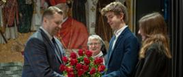 Minister Czarnek w marynarce w kratę stoi przed młodym mężczyzną w garniturze i trzyma bukiet czerwonych kwiatów, obok stoi młoda kobieta ubrana na czarno, za nimi zakonnica w szarym ubraniu.