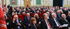 Widok na wypełnioną siedzącymi na krzesłach ludźmi salę, za nimi na biało-zielonych ścianach wiszą portrety.