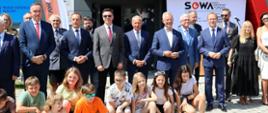 Na dworze w ostrym słońcu przed wejściem do szarego budynku z napisem SOWA stoi grupa ludzi, przed nimi na ziemi siedzą dzieci.