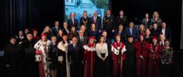 Zdjęcie zbiorowe, na podwyższeniu stoi duża grupa ludzi, w pierwszym rzędzie kadra uczelni w ceremonialnych strojach - czerwonych płaszczach z gronostajowymi kołnierzami.