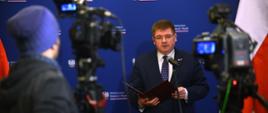 Wiceminister stoi i trzyma w ręku teczkę z orłem, mówi do mikrofonu na stojaku, za nim niebieska ściana, a przed nim kilka kamer na statywach i dziennikarz w niebieskiej czapce.