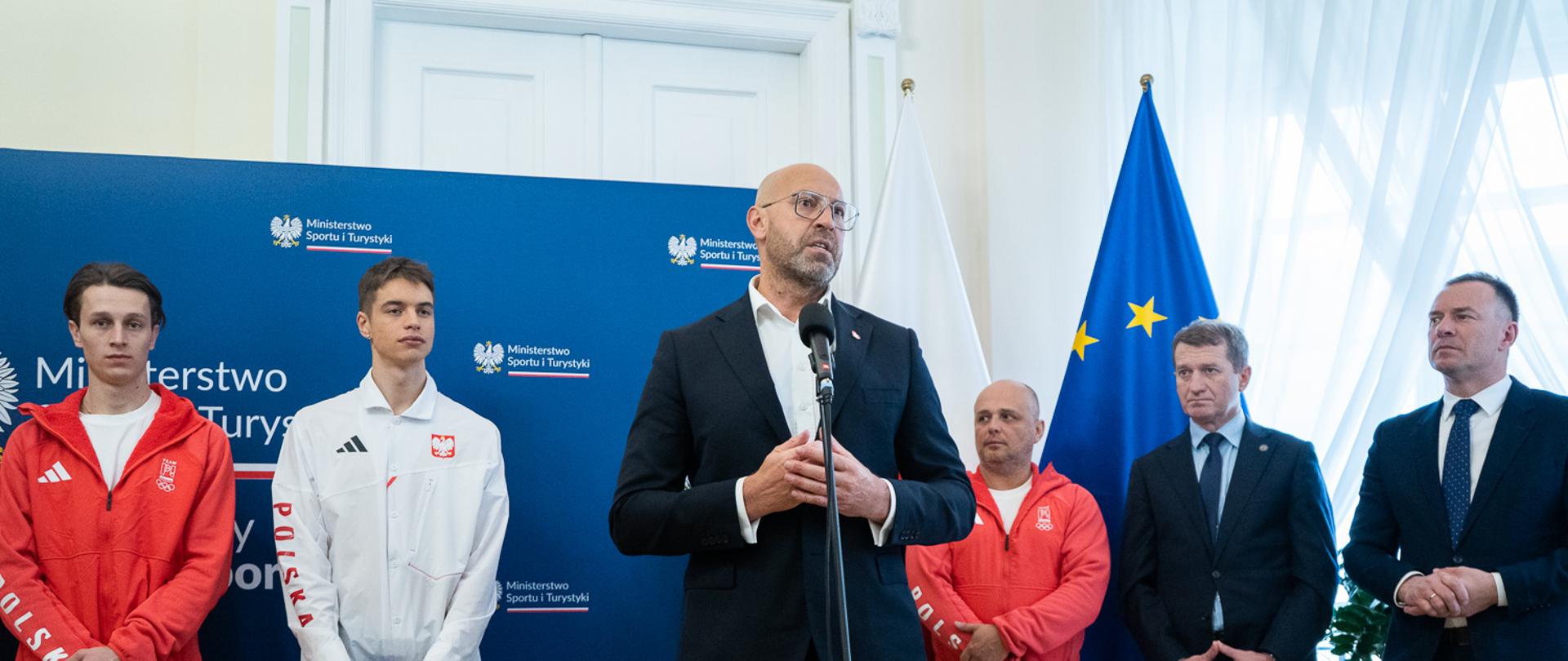 Spotkanie ministra z medalistami igrzysk olimpijskich. Minister stoi za mikrofonem na tle granatowej ścianki z logiem ministerstwa. Po prawej stronie w garniturach stoją sekretarze stanu a po lewej stronie sportowcy w białych i czerwonych ubraniach