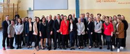 Zdjęcie zbiorowe, na sali pod ścianą stoi duża grupa ludzi. pomiędzy nimi wiceminister Katarzyna Lubnauer. W tle widać drabinki gimnastyczne i napis na ścianie: Życie ludzkie to olbrzymie zawody sportowe, których jesteśmy zarówno uczestnikami jak i widzami... Antoni Golubiew 