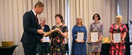 Wiceminister Piontkowski wręcza kobiecie w sukience w kolorowe kwiaty oprawiony w ramki dyplom, za nimi stoi grupa kobiet trzymających dyplomy.