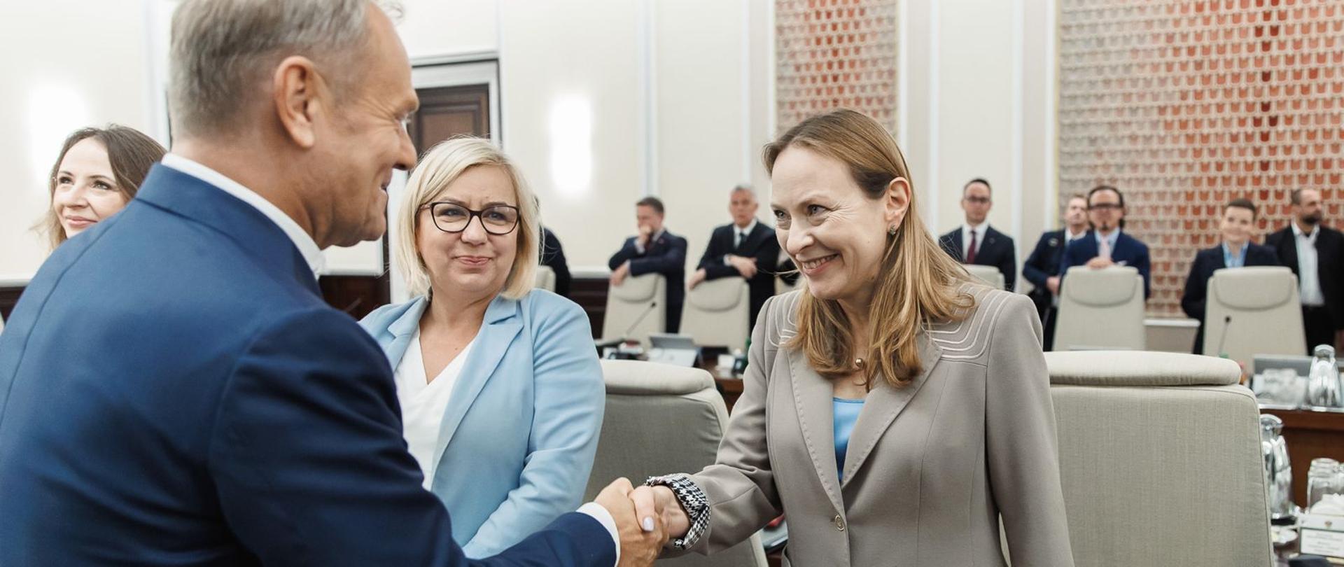 Dwie osoby stoją na przeciwko siebie i podają sobie dłonie. Z lewej strony odwrócony bokiem stoi premier Donald Tusk, a z prawej strony uśmiechnięta stoi minister Katarzyna Pełczyńska-Nałęcz.