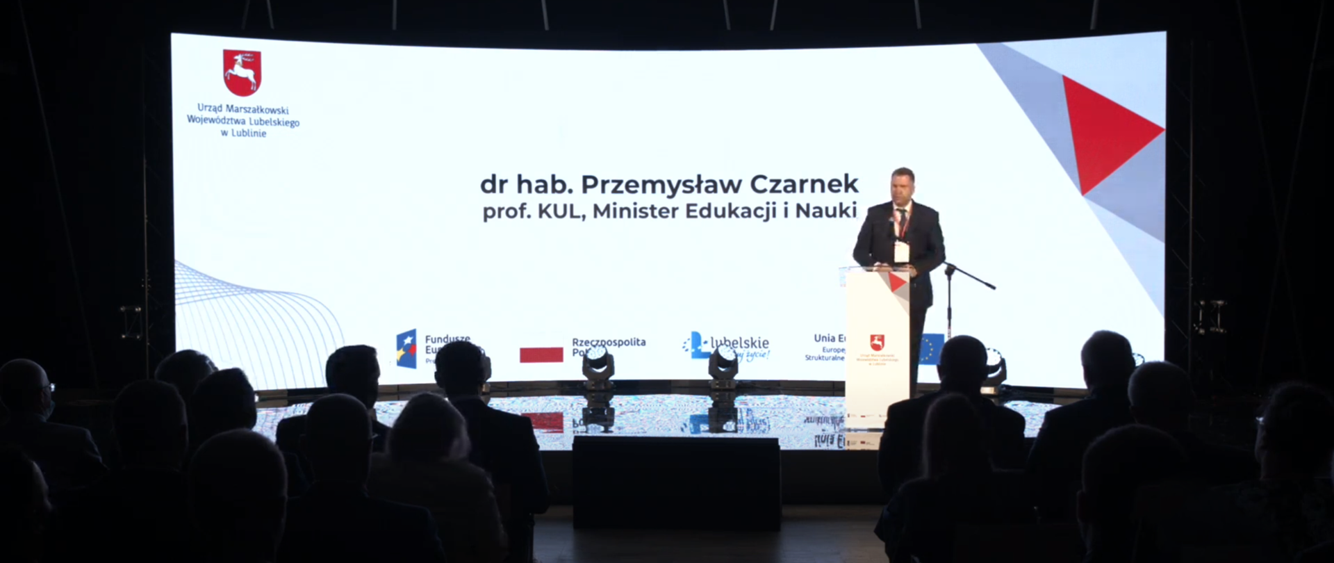 Sala konferencyjna. widownia siedzi w cieniu, w tle ekran informujący o osobie przemawiającej przy pulpicie – ministrze edukacji i nauki Przemysławie Czarnku