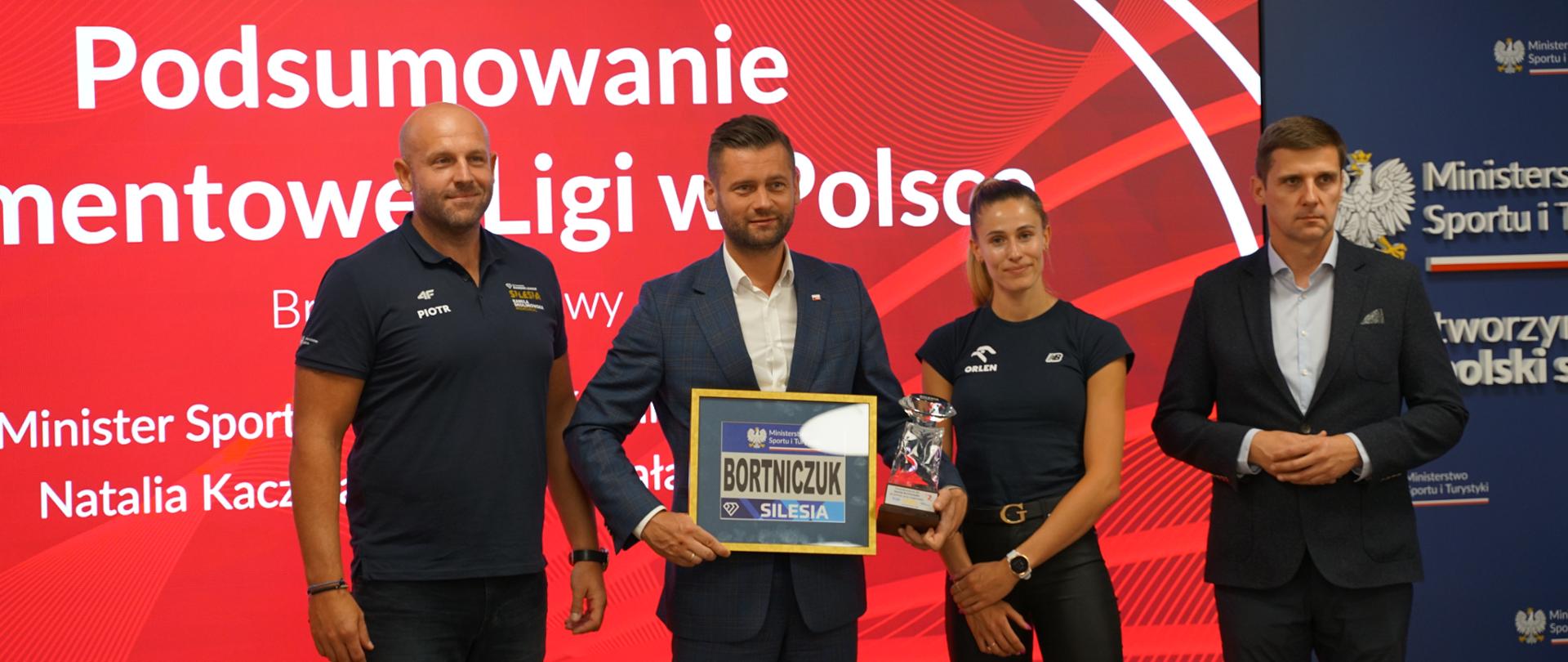 Minister Kamil Bortniczuk prezentuje otrzymane od organizatorów Memoriału Kamili Skolimowskiej upominki - okolicznościową szklaną statuetkę oraz numer startowy z nazwiskiem Bortniczuk w ramce za szkłem. Razem z ministrem stoją Piotr Małachowski - dyrektor mityngu, Natalia Kaczmarek - biegaczka i sekretarz generalny PZLA Krzysztof Kęcki.