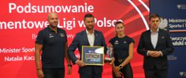 Minister Kamil Bortniczuk prezentuje otrzymane od organizatorów Memoriału Kamili Skolimowskiej upominki - okolicznościową szklaną statuetkę oraz numer startowy z nazwiskiem Bortniczuk w ramce za szkłem. Razem z ministrem stoją Piotr Małachowski - dyrektor mityngu, Natalia Kaczmarek - biegaczka i sekretarz generalny PZLA Krzysztof Kęcki.