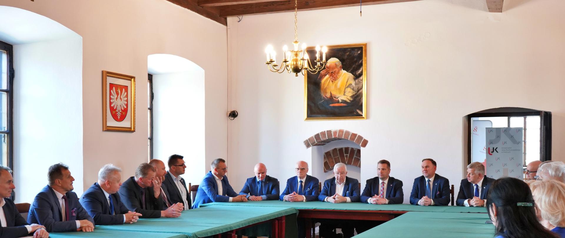 W sali z białymi ścianami stoją ustawione w czworokąt drewniane stoły przykryte zielonym materiałem, przy nich siedzi duża grupa elegancko ubranych osób, z lewej dwa duże okna, między którymi wisi godło.
