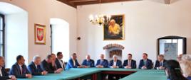W sali z białymi ścianami stoją ustawione w czworokąt drewniane stoły przykryte zielonym materiałem, przy nich siedzi duża grupa elegancko ubranych osób, z lewej dwa duże okna, między którymi wisi godło.