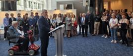 Widok na podwórko pomiędzy białymi budynkami, wiceminister Murdzek stoi i mówi do mikrofonu na stojaku, za nim mężczyzna na czterokołowym wózku inwalidzkim, za nimi grupa ludzi.