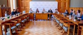 W sali stoją stoły i krzesła ustawione w formie odwróconej litery "u". Pośrodku siedzi wiceminister Paulina Piechna-Więckiewicz i zabiera głos. Za jej plecami stoi ścianka z logiem Ministerstwa Edukacji Narodowej. Przy pozostałych stołach siedzą uczestnicy spotkania. 