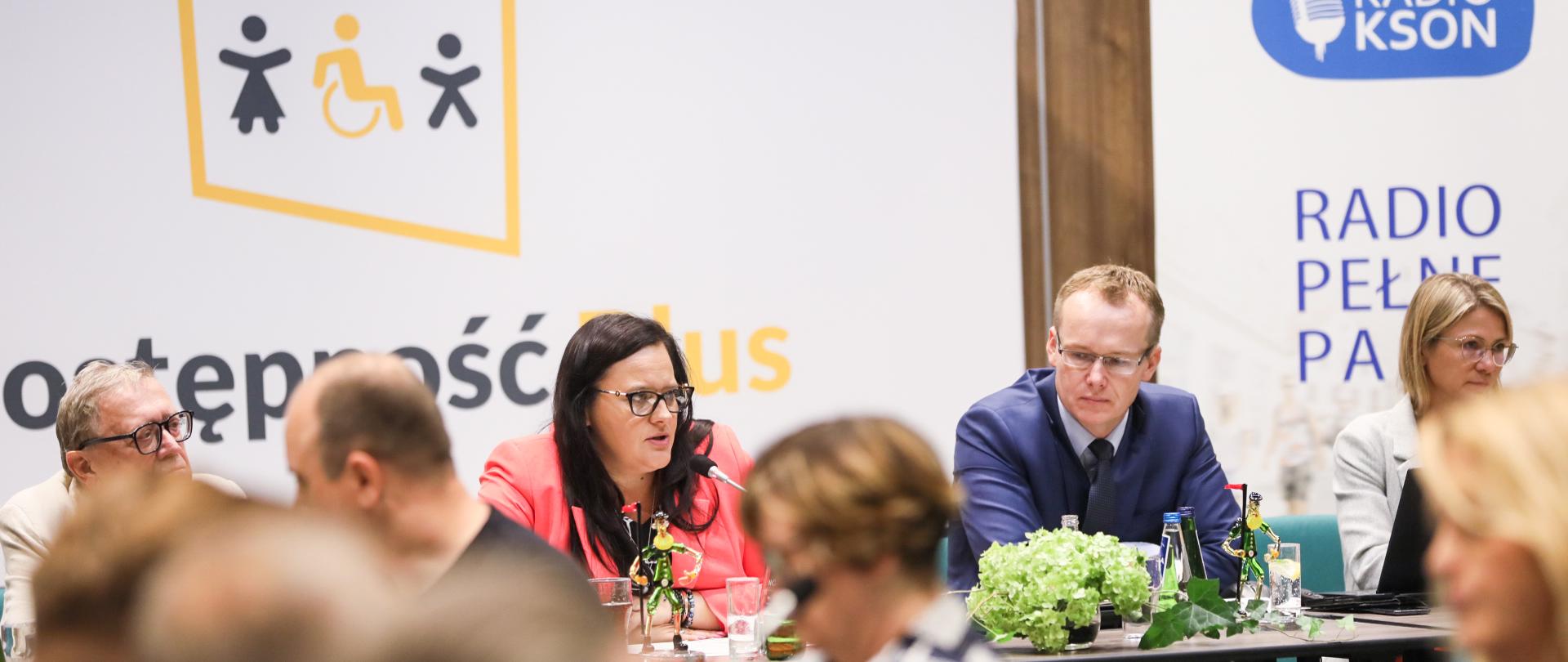 Posiedzenie Rady Dostępności. Na pierwszym planie niewyraźne sylwetki osób. Za nimi przy stole konferencyjnym siedzą cztery osoby.