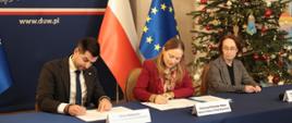 Na zdjęciu minister Katarzyna Pełczyńska-Nałęcz siedzi za stołem konferencyjnym i podpisuje dokumenty w towarzystwie innych uczestników spotkania
