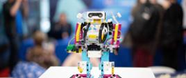 Na białym stoliku stoi mały robot złożony z kolorowych elementów, w tle sala z ludźmi.