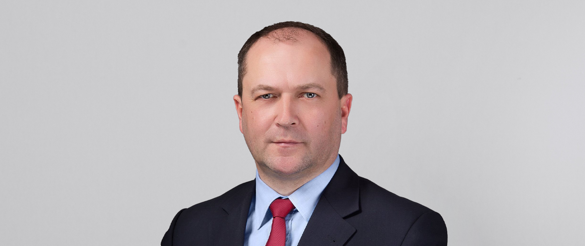 Na tle jasnoszarej ściany stoi wiceminister Kiepura w czarnym garniturze.