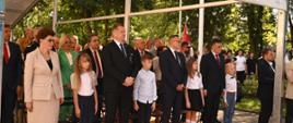 Pod namiotowym daszkiem stoi minister Czarnek, kilka innych osób i galowo ubrane dzieci, za nimi drzewa.