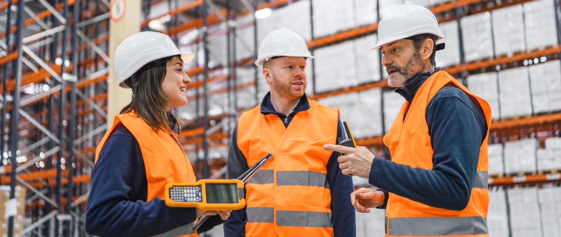 Warehouse workers wearing safety vests and hardhats are discussing logistics and using handheld devices in a large warehouse or distribution center