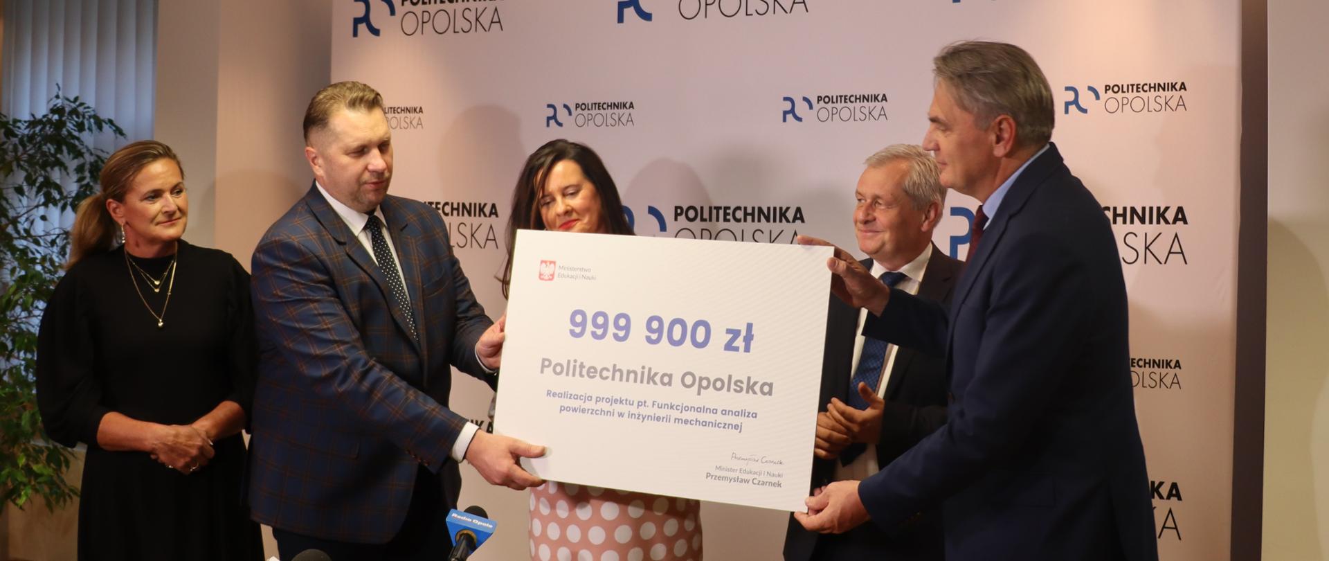 Grupa osób, trzech mężczyzn i dwie kobiety. Minister Przemysław Czarnek wręcza czek z kwotą 999900 mężczyźnie w garniturze na tle ścianki z napisami Politechnika Opolska 
