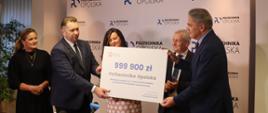 Grupa osób, trzech mężczyzn i dwie kobiety. Minister Przemysław Czarnek wręcza czek z kwotą 999900 mężczyźnie w garniturze na tle ścianki z napisami Politechnika Opolska 