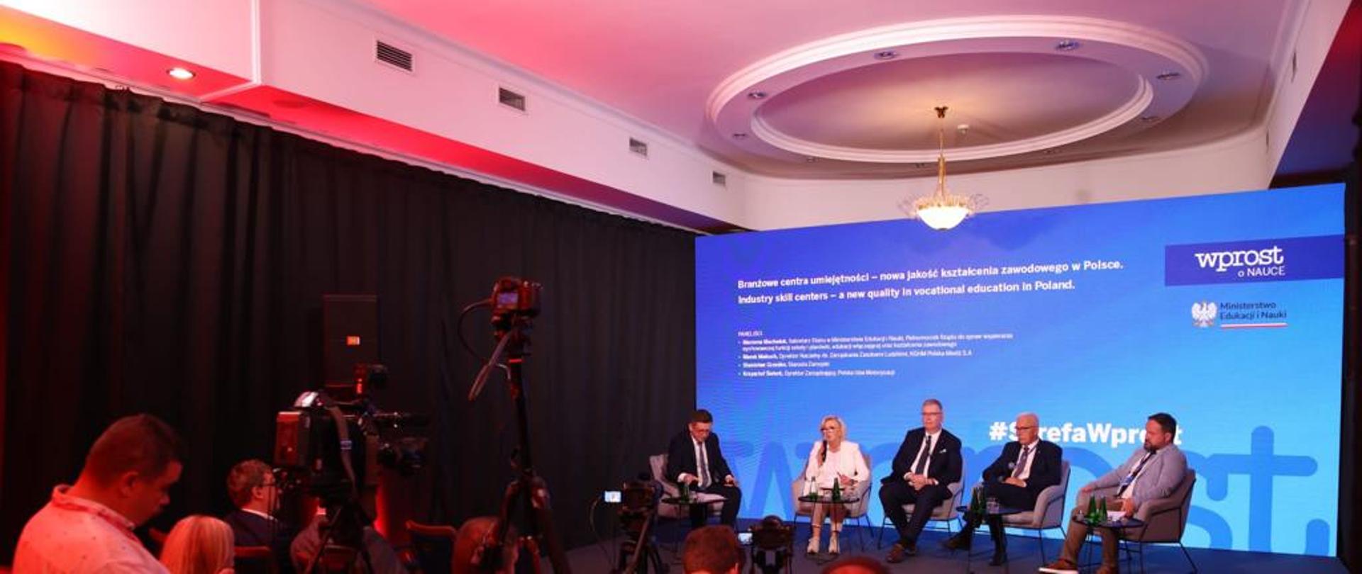 Pod niebieską ścianą siedzi na krześle wiceminister Machałek i kilku mężczyzn w garniturach, naprzeciwko sala w której ludzie siedzą na krzesłach.