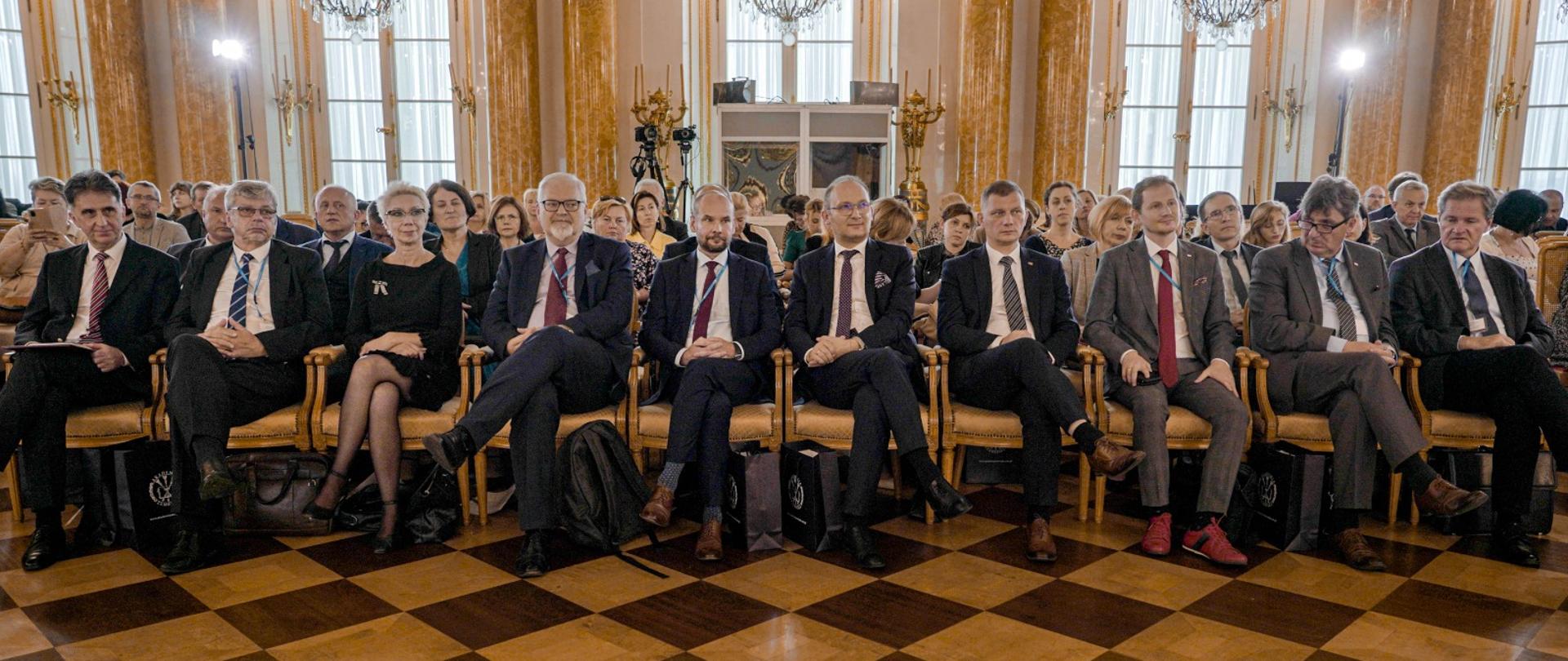 Zdjęcie od przodu, w eleganckiej sali ze zdobionymi złoceniami ścianami na podłodze z żółtych i brązowych czworokątnych płyt stoją ustawione w rzędy krzesła, na nich siedzi dużo ludzi.