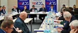Widok na salę z żółtymi ścianami, przy ustawionych w czworokąt stołach siedzą ludzie, u szczytu stołu siedzi minister Czarnek, kobieta w niebieskiej marynarce i dwóch mężczyzn w garniturach, za nimi banery z napisem WRDS i zasłonięte żaluzjami okna.
