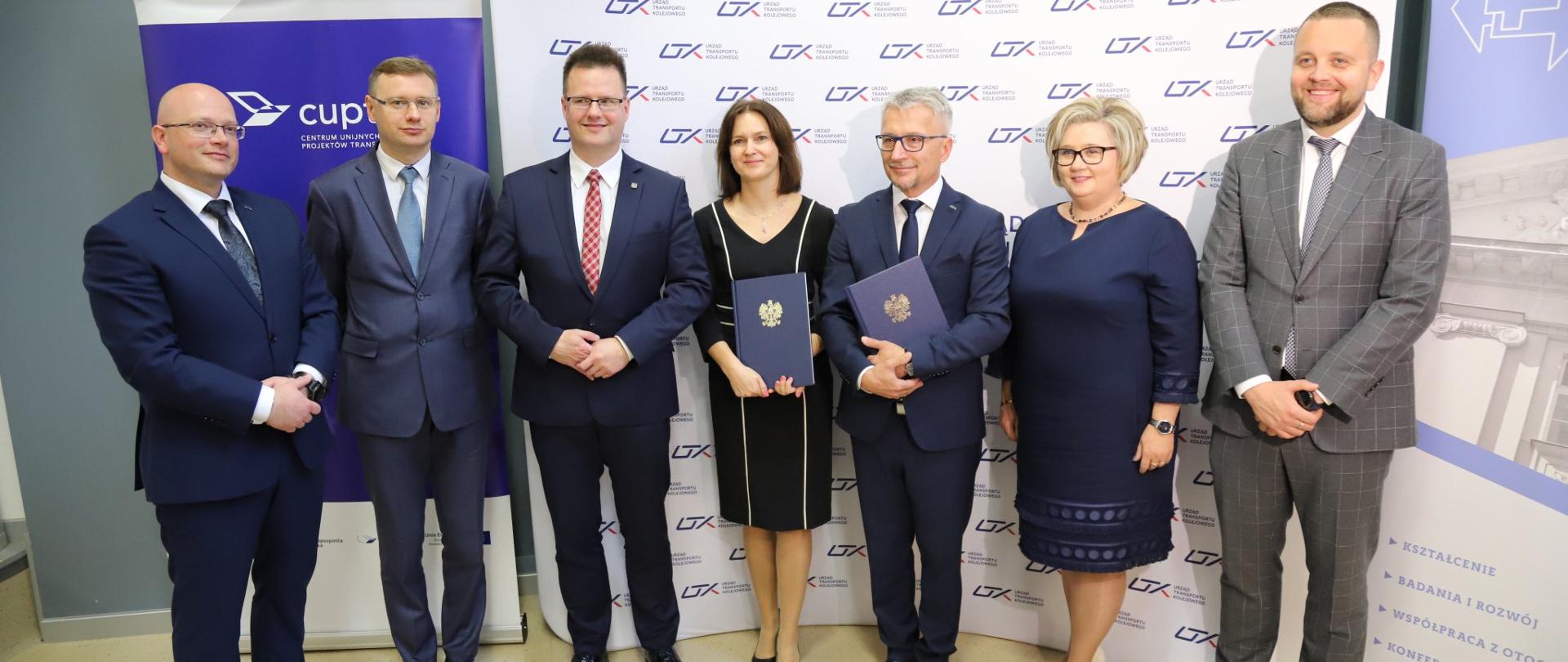 Na tle banerów UTK i CUPT stoi grupa ludzi - 5 mężczyzn i 2 kobiety. Wśród nich Joanna Lech, Dyrektora Centrum Unijnych Projektów Transportowych oraz Ignacy Góra, Prezes Urzędu Transportu Kolejowego.