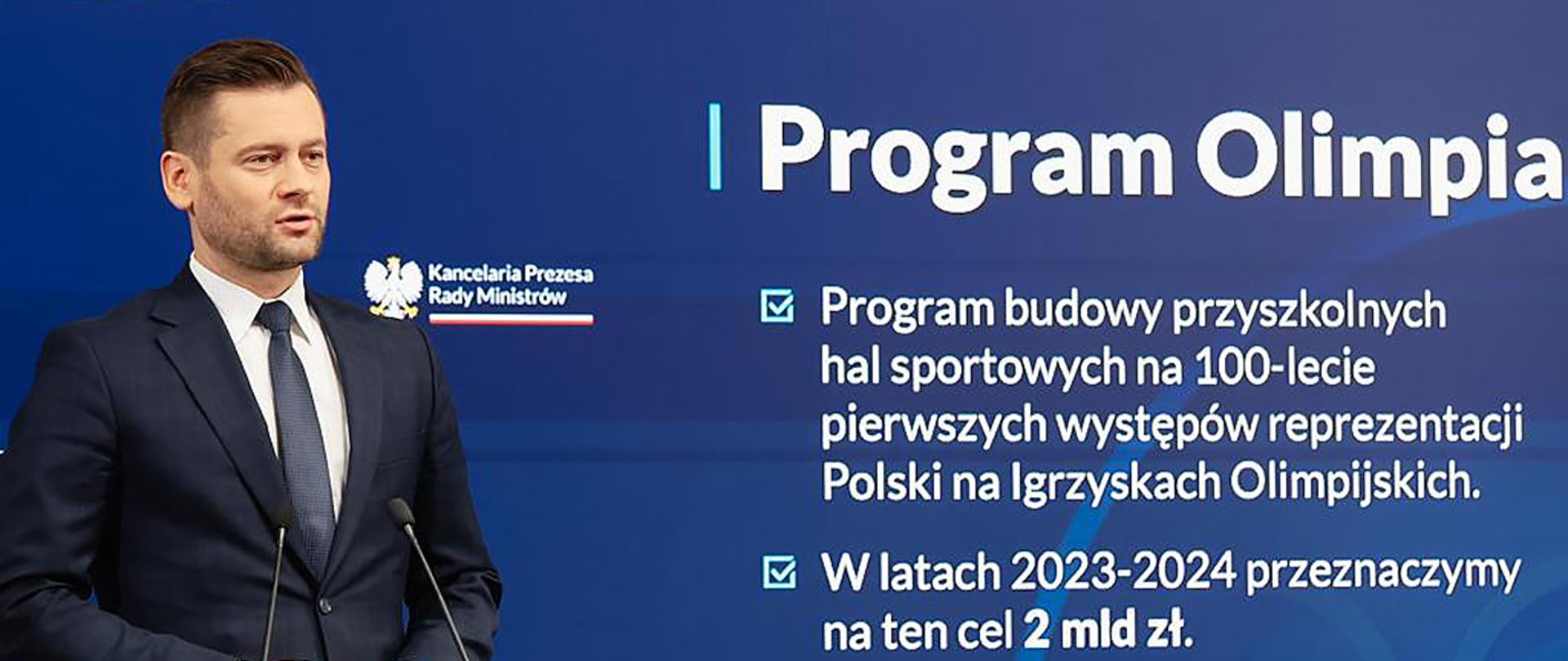 OLIMPIA - program przyszkolnych hal sportowych - minister Kamil Bortniczuk podczas konferencji w KPRM dotyczącej rozpoczęcia programu; minister stoi na tle ekranu z wyświetlonymi informacjami o programie