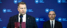 Minister Czarnek stoi obok mężczyzny w garniturze, trzyma w ręku fioletową teczkę, za nimi niebieska ściana.