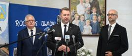 Minister Czarnek stoi pod ścianą na której wisi rodzinny portret, obok niego dwóch mężczyzn w garniturach, przed nimi kilka mikrofonów.