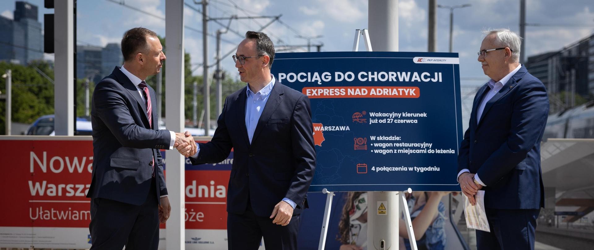 Piotr Borys na konferencji na dworcu kolejowym. Na pierwszym planie stoi trzech mężczyzn, za nimi niebieska tablica oraz tory kolejowe
