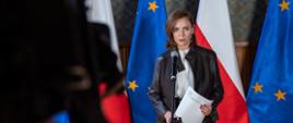 Wiceminister Joanna Mucha stoi przy mikrofonie w ręku trzyma notatki. W tle flagi biało-czerwone i niebieskie ze złotymi gwiazdkami. 