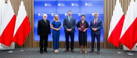 Pod ścianą stoi grupa pięciu osób, pośrodku minister Czarnek, stojąca obok niego kobieta w czarnym ubraniu trzyma fioletową teczkę z godłem, po bokach grupy z obu stron polskie flagi.