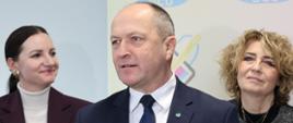Minister Henryk Kiepura, sekretarz stanu w MEN, w pozycji stojącej udziela wypowiedzi dla mediów. Minister ma na sobie granatowy garnitur, białą koszulę i granatowy krawat. Przed nim są trzy mikrofony stacji radiowych i telewizyjnych. Obok ministra stoją dwie kobiety, w tym Hanna Zdanowska, prezydent Łodzi. Za postaciami widać wyświetlany napis "BCU" i logo centrum.