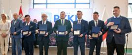 W hali sportowej stoi szereg mężczyzn w garniturach, trzymają w rekach niebieskie otwarte pudełka, w których leżą dokumenty, dalej biała ściana i duże okno.