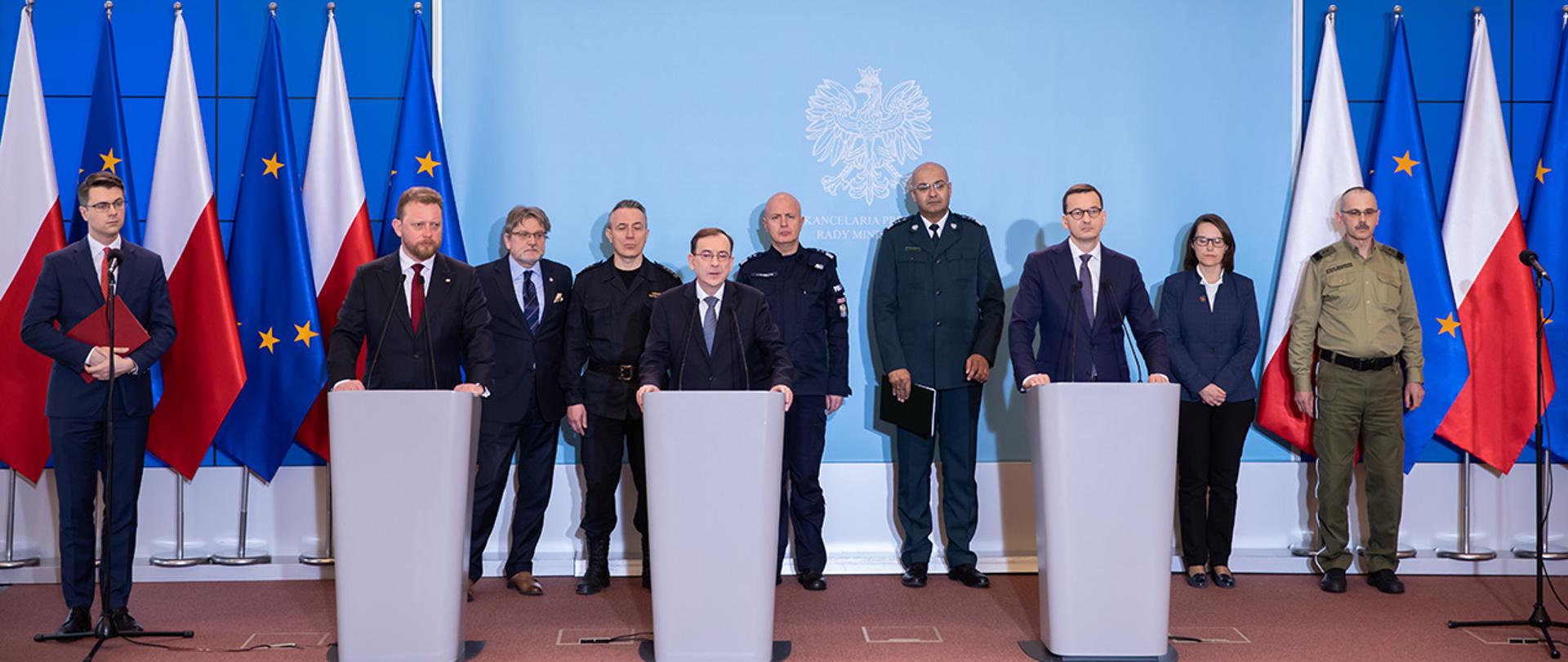 Ma zdjęciu widać m.in. premiera Mateusza Morawieckiego, ministra Mariusza Kamińskiego oraz ministra Łukasza Szumowskiego stojących przy mównicach w trakcie konferencji prasowej w KPRM. W tle stoja m.in. nadbryg. ANDRZEJ BARTKOWIAK komendant PSP, gen. insp. Jarosław Szymczyk komendant główny Policji i gen. dyw. SG Tomasz Praga komendant główny Straży Granicznej. W tle widać również dużą planszę z orłem z godła narodowego napisem " Kancelaria Prezesa Rady Ministrów i flagi Polski i UE.