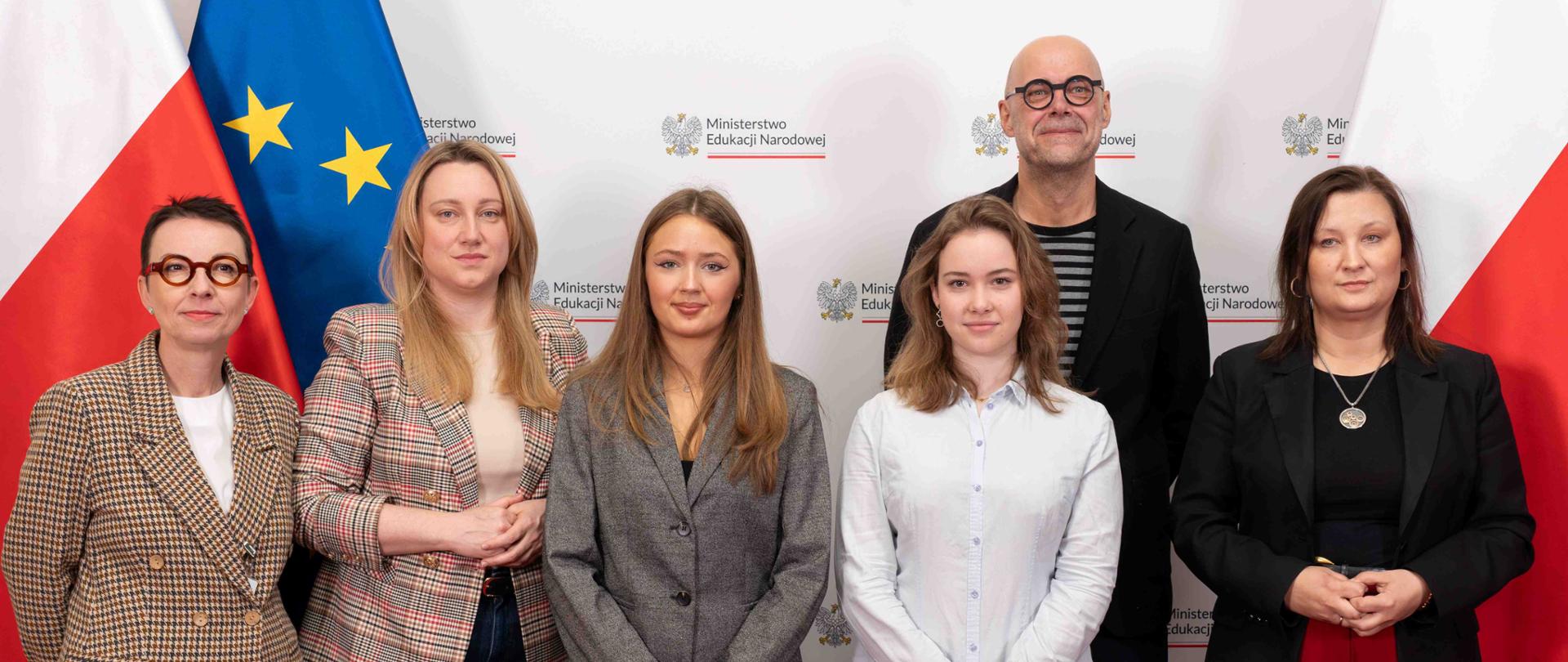 Na tle białej ścianki z napisem Ministerstwo Edukacji Narodowej stoi wiceminister Paulina Piechna-Więckiewicz, obok niej grupa osób. W tle flagi Polski i UE.