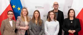 Na tle białej ścianki z napisem Ministerstwo Edukacji Narodowej stoi wiceminister Paulina Piechna-Więckiewicz, obok niej grupa osób. W tle flagi Polski i UE.