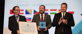 Minister Czarnek i dwóch mężczyzn w garniturach z których jeden trzyma duży dokument w ramkach a drugi czworokątną statuetkę stoją pod ekranem, na którym widać obrazki różnych flag.