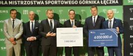Pod zieloną ścianą stoi grupa sześciu mężczyzn, jeden pośrodku trzyma wielki symboliczny czek z napisem 3 000 000 zł, dwóch stojących z prawej razem trzyma niebieską tabliczkę z napisem 6 050 000 zł.