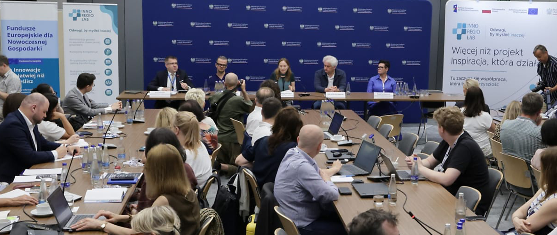 Sala konferencyjna podczas panelu dyskusyjnego. Przy długim stole siedzi sześcioro prelegentów, a przed nimi zgromadzona publiczność z laptopami i notatkami. W tle widoczne są banery programu InnoRegioLab oraz Funduszy Europejskich, a jeden z uczestników wykonuje zdjęcia panelu.