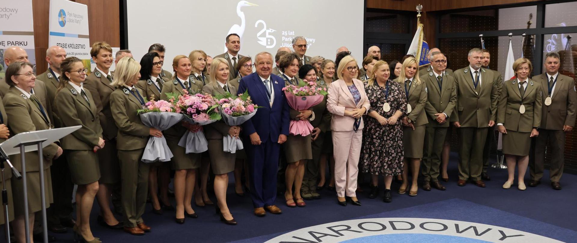 Ministra klimatu i środowiska Paulina Hennig-Kloska podczas jubileuszowej konferencji na terenie Parku Narodowego „Ujście Warty”. Ministra stoi wraz z innymi uczestnikami konferencji. Wszyscy wspólnie pozują do zdjęcia grupowego.