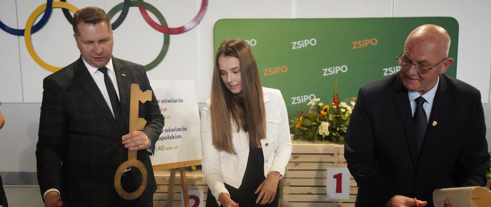 W sali na tle zielonej ścianki z napisem ZSiPO stoi minister Czarnek trzymający w ręku duży symboliczny złoty klucz, kobieta w białej marynarce i mężczyzna w czarnym garniturze, wspólnie przecinają rozciągniętą przed nimi białą wstęgę.