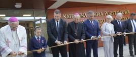 Grupa kilku osób stoi przed żółtą wstęgą, każdy trzyma nożyczki i równocześnie wszyscy przecinają wstęgę.