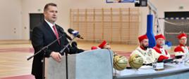 Na środku sali gimnastycznej za szarą mównicą stoi minister Czarnek i mówi do dwóch mikrofonów, za nim za szarym stolikiem siedzi kilku mężczyzn w czerwonych ceremonialnych strojach z gronostajowymi kołnierzami.