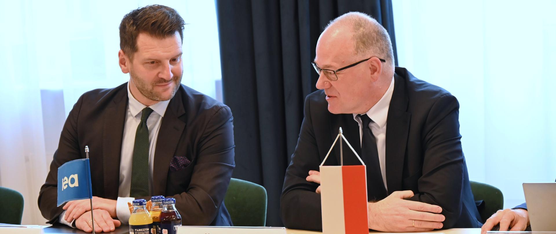Na zdjęciu widoczne są dwie osoby. Za stołem po lewej stronie siedzi Miłosz Karpiński z International Energy Agency, a po prawej Krzysztof Galos, Podsekretarz Stanu w MKiŚ, Główny Geolog Kraju. Na stole stoją dwie niewielkie flagi, flaga Polski i flaga International Energy Agency.