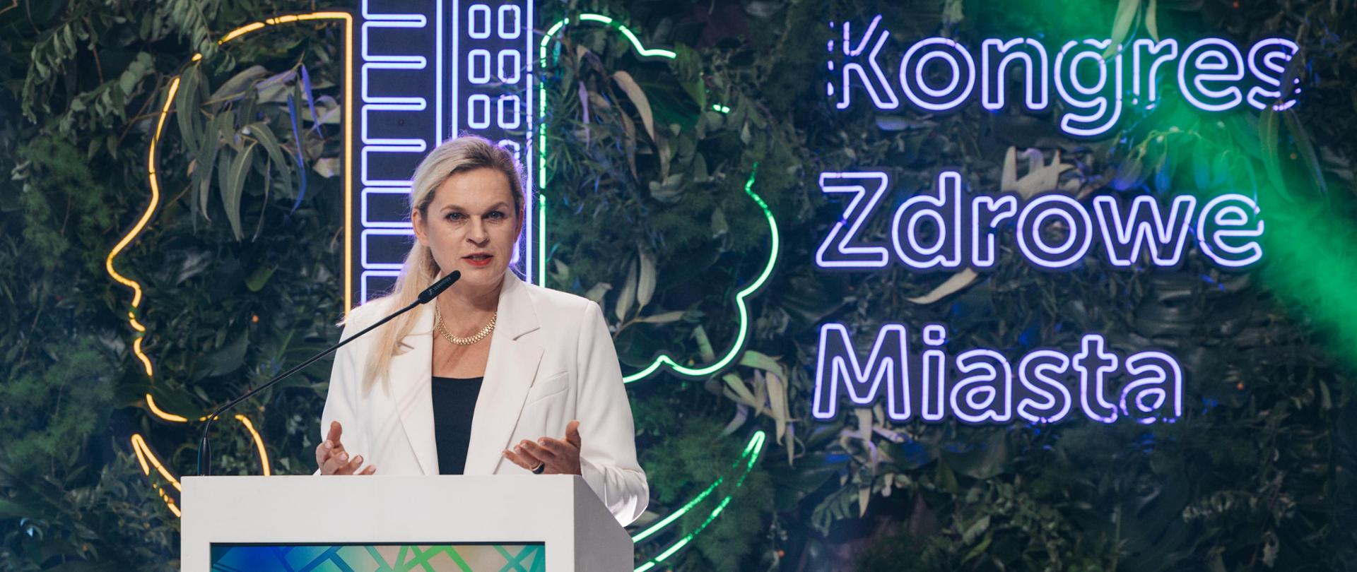 Barbara Nowacka, Minister Edukacji, stoi za białą mównicą i przemawia. Ma na sobie białą marynarkę i czarną bluzkę. W tle neonowy napis "Kongres Zdrowe Miasta" jest otoczony bujną, zieloną roślinnością i neonowymi konturami głowy oraz wieżowca.