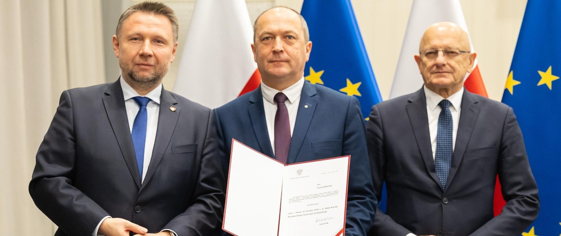 Trzech mężczyzn w garniturach, mężczyzna w środku trzyma otwarty dokument. W tle flagi biało-czerwone i niebieskie z żółtymi gwiazdkami