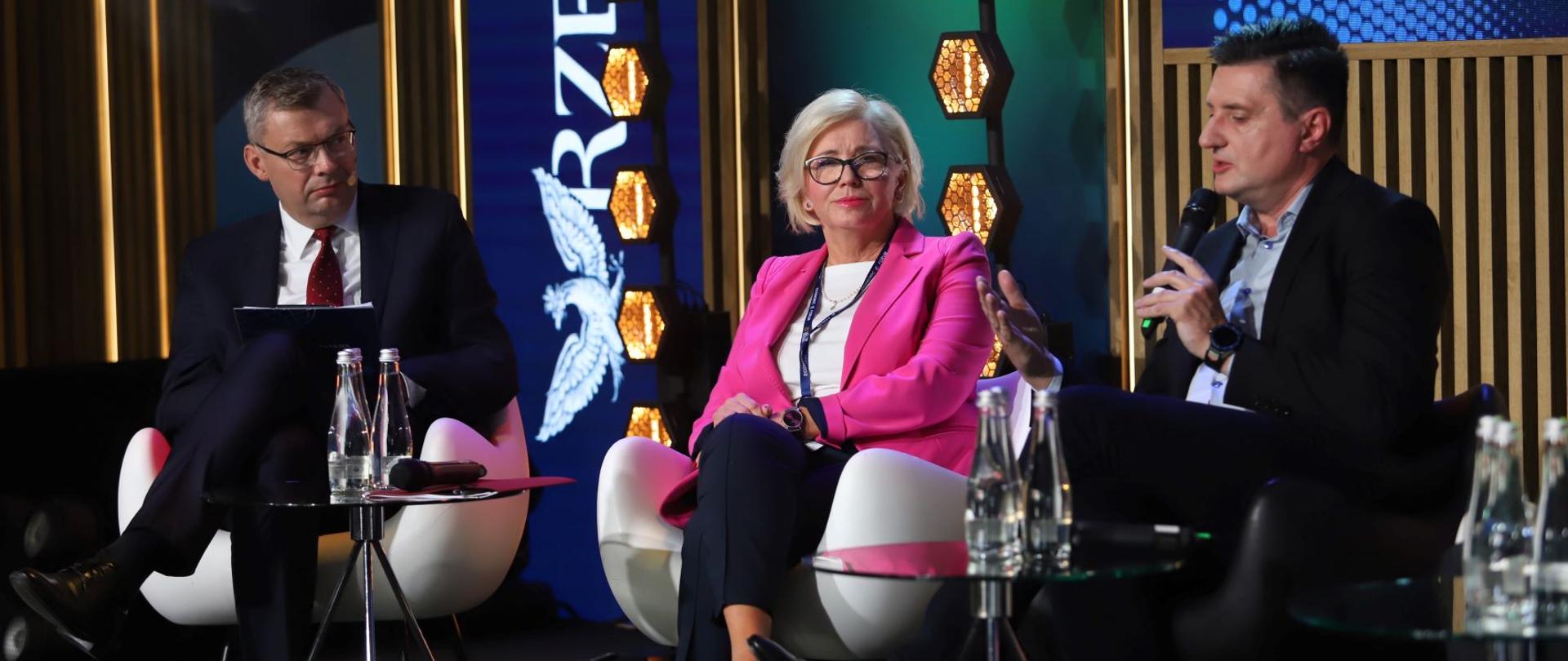 Pod kolorową ścianą siedzi wiceminister Machałek w różowym ubraniu, po jej obu stronach mężczyźni w garniturach.